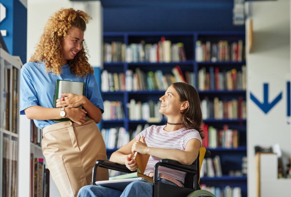Vertical full length portrait of smiling young woman with disability talking to friend in college library