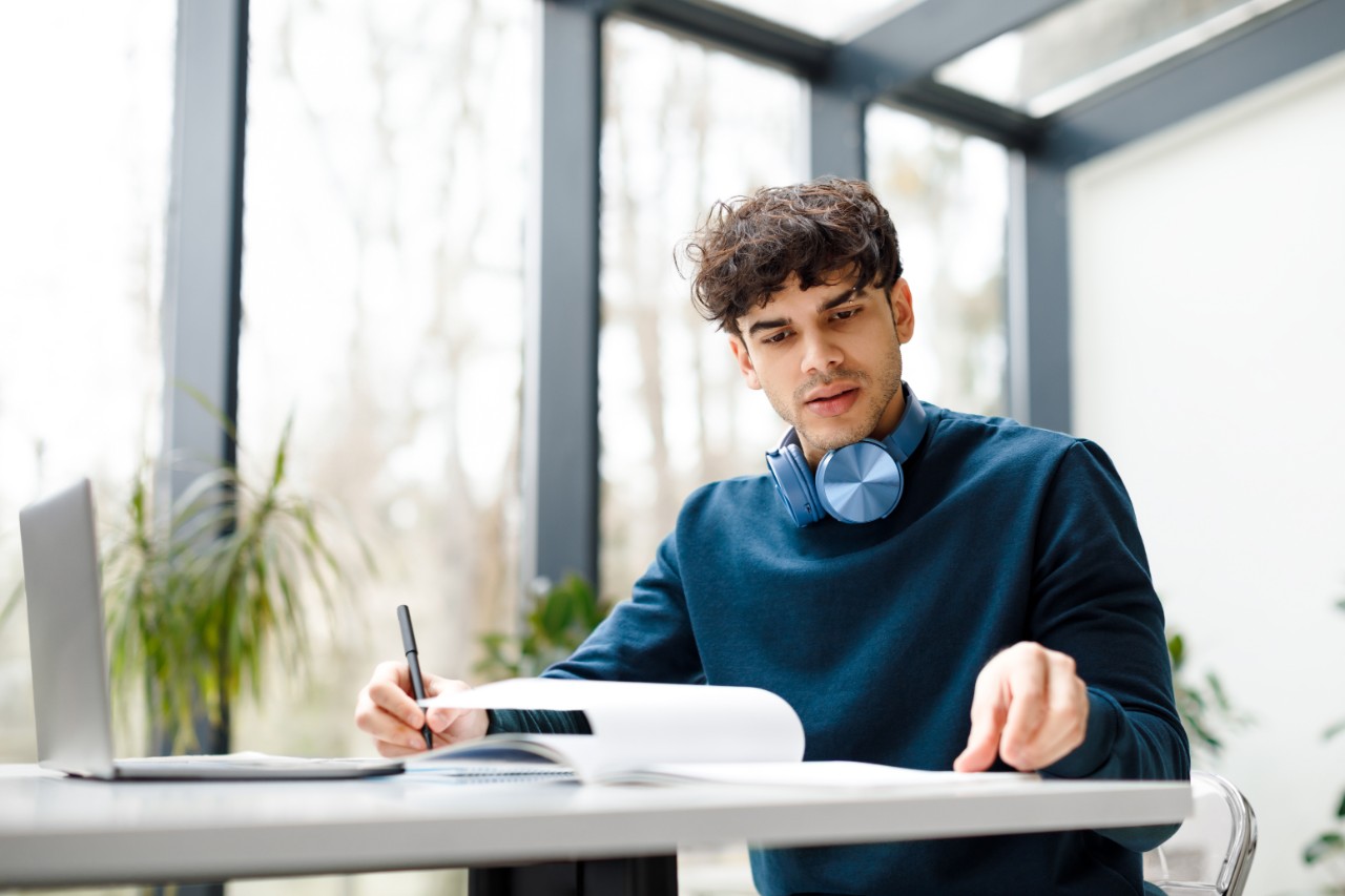 European student guy having online lesson on laptop, writing down info while participating in webinar, learning remotely, sitting in coworking space, copy space