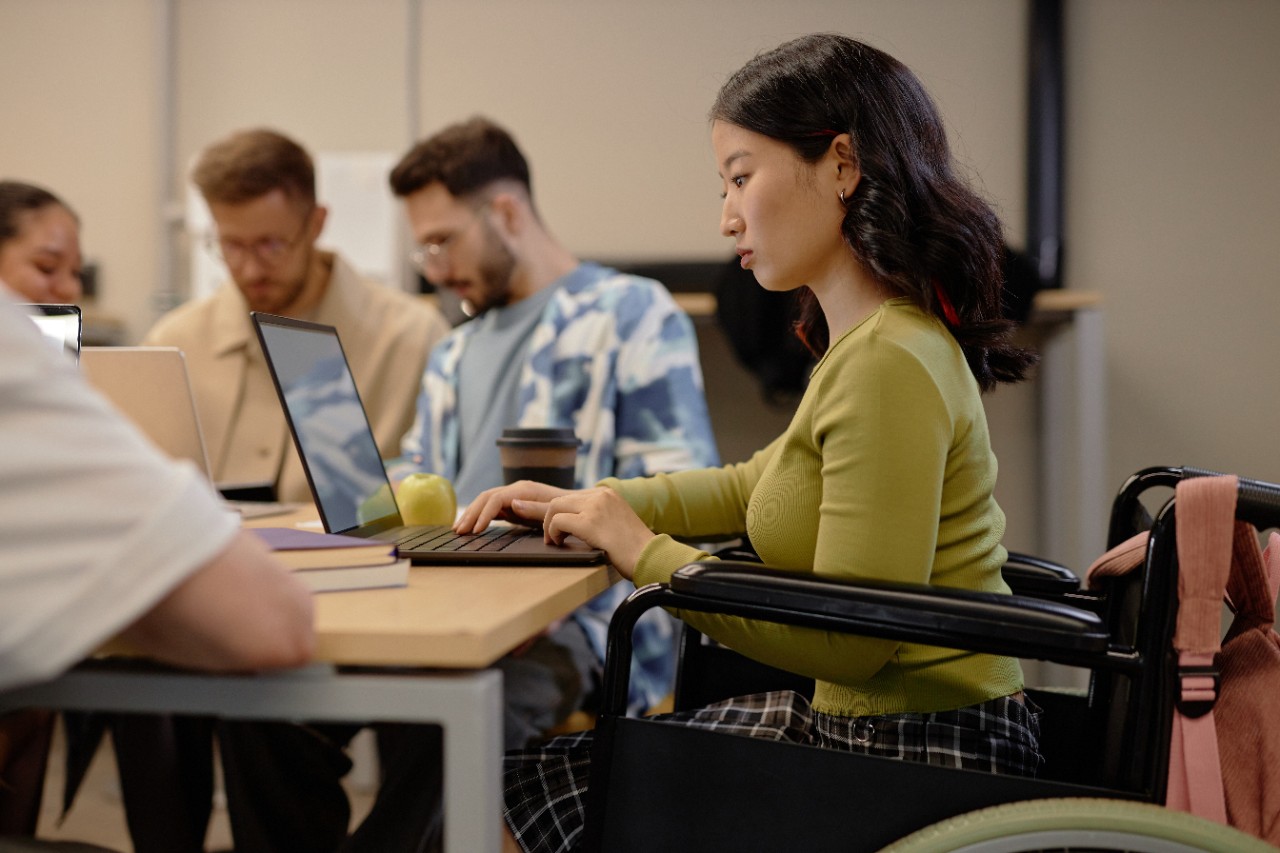 Side view of focused Asian female wheelchair user working on laptop at university group seminar