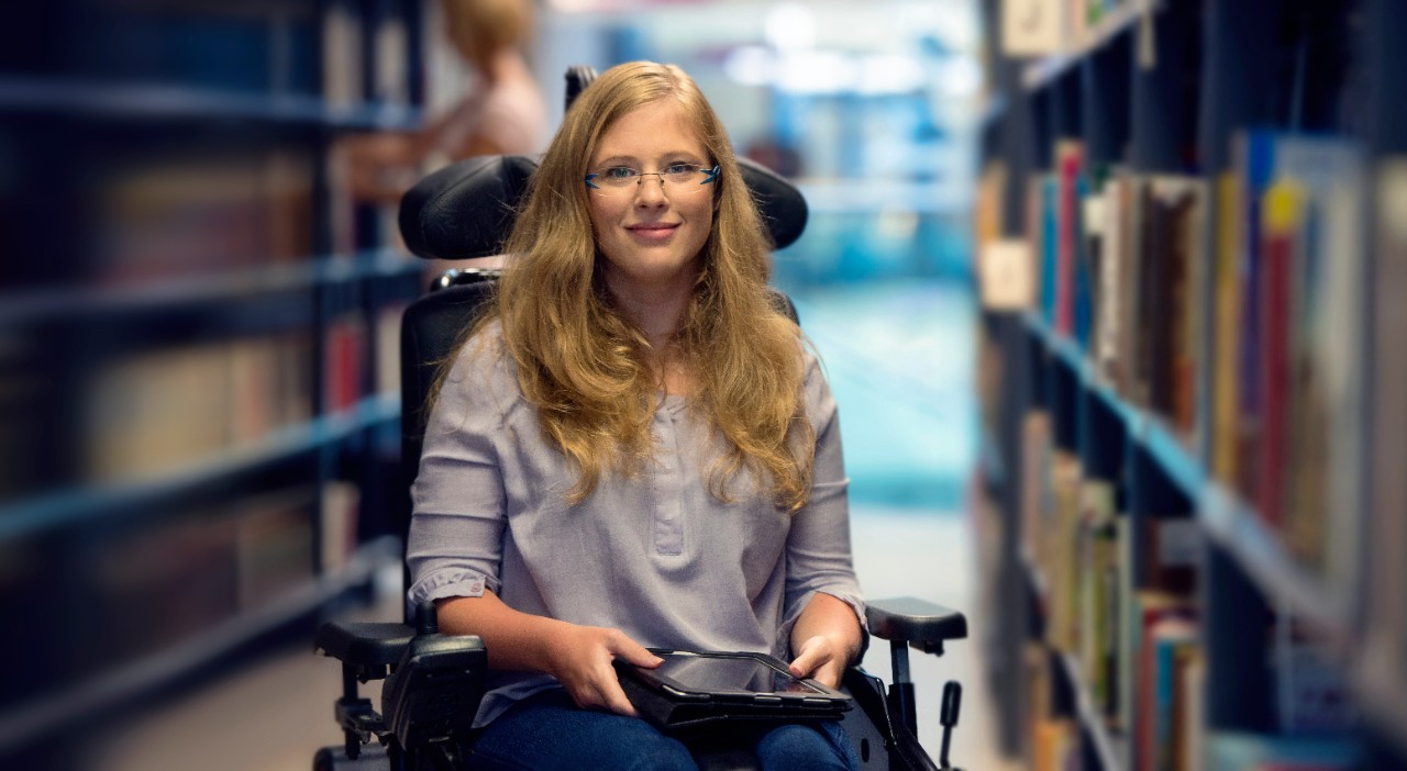 Young woman sitting in wheelchair holding digital tablet, looking at camera and smiling.