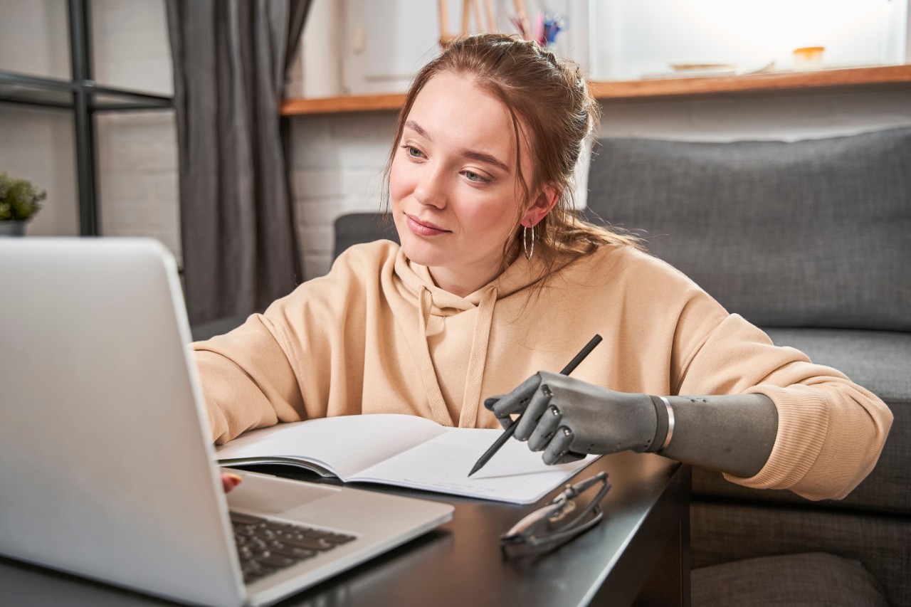 Beautiful young woman with prosthesis arm writing at notebook while sitting at the table with her laptop computer. Adorable girl looking at the screen while working at home